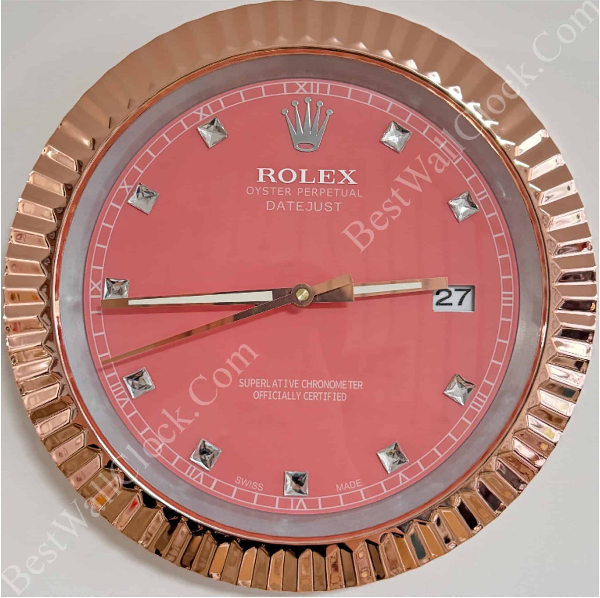 Close-up of a round wall clock styled like a Rolex Datejust watch with a rose gold fluted bezel, pink dial, diamond-shaped hour markers, and date display showing the 27th.