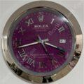 A Rolex Oyster Perpetual Datejust wall clock with a purple face, silver Roman numerals, silver hour and minute hands, a date display showing 27, and a reflective silver frame.