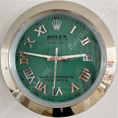 Round Rolex wall clock with green face, silver Roman numerals, silver frame, and date display showing 18, with hour hand near 3, minute hand near 9, and second hand near 6.