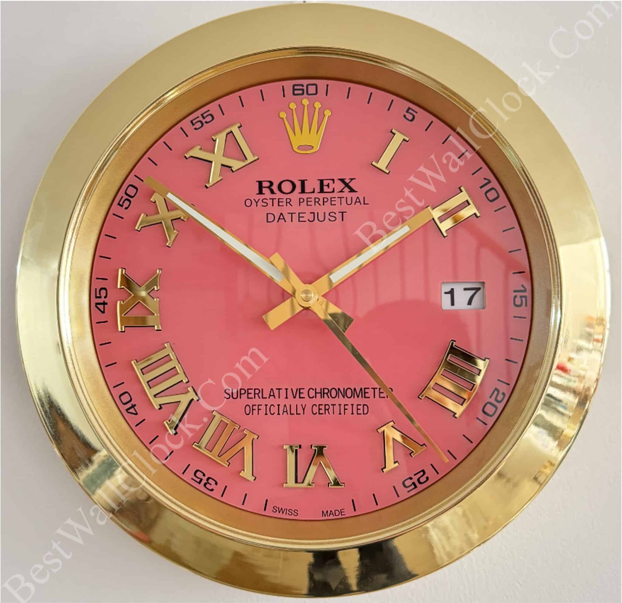 Round wall clock with a gold frame and a pink face featuring gold Roman numerals, three gold hands, a date window showing 17, and Rolex branding including the crown logo and text.