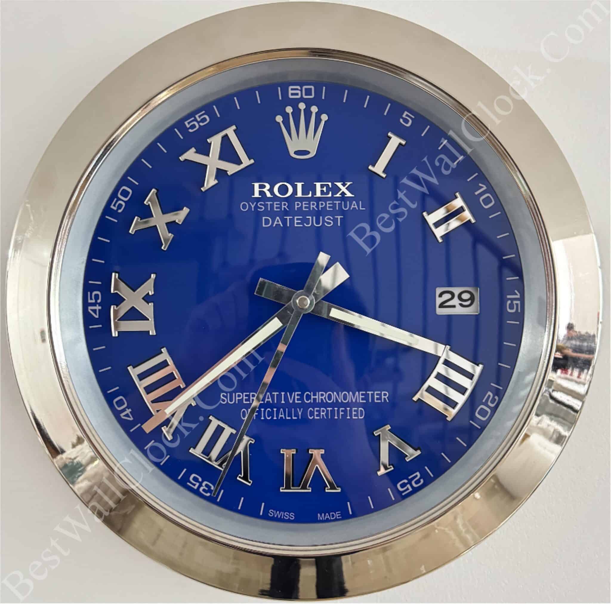 Close-up of a round Rolex wall clock with a blue face, silver Roman numeral hour markers, and a silver frame, showing the time as 8:19:35 with a date window displaying the number 29 and text indicating it is an Oyster Perpetual Datejust model.