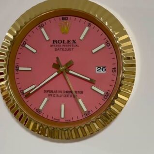 Close-up of a gold-framed Rolex wall clock with a pink face, showing the time as approximately 3:39 and the date as 26.