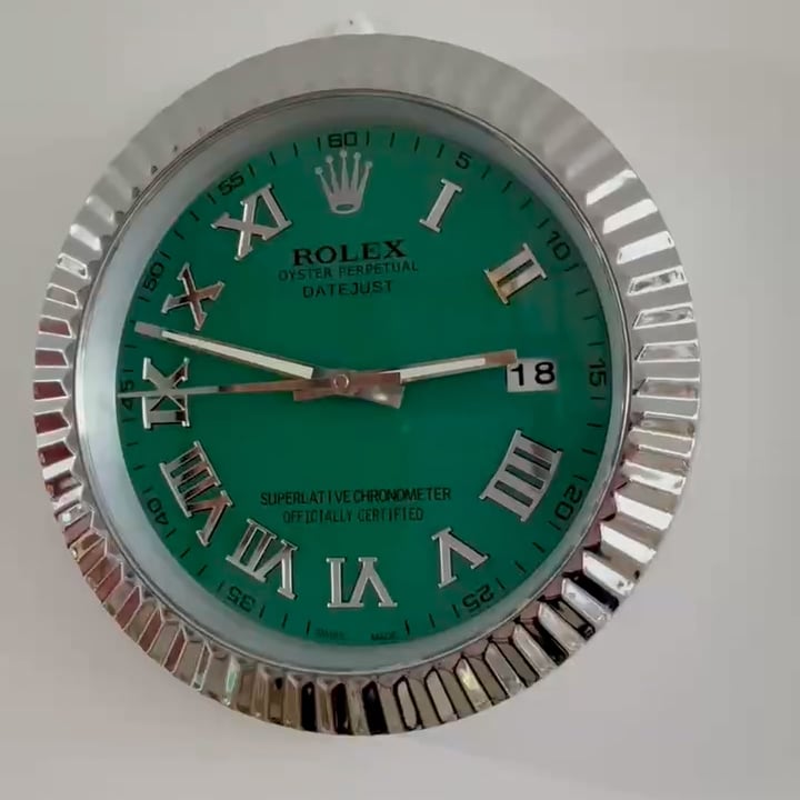 Close-up of a Rolex Oyster Perpetual Datejust wall clock with a green face, silver Roman numerals, and a date display showing the 18th, set in a reflective silver fluted bezel.