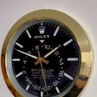 Close-up of a circular Rolex Sky-Dweller wall clock with a black dial, white hour markers, gold hands, and a gold bezel set against a light background.