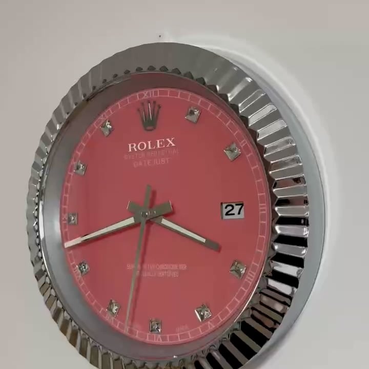 Close-up of a wall clock styled like a Rolex watch with a red face, silver rim, and diamond-like hour markers showing the time as 3:42:33 and the date as the 27th.