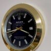 Rolex Oyster Perpetual Day-Date wall clock with black face, gold rim, diamond hour markers, showing Tuesday and date 17, mounted on a white wall