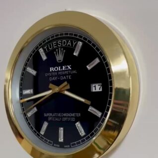 Rolex Oyster Perpetual Day-Date wall clock with black face, gold rim, diamond hour markers, showing Tuesday and date 17, mounted on a white wall