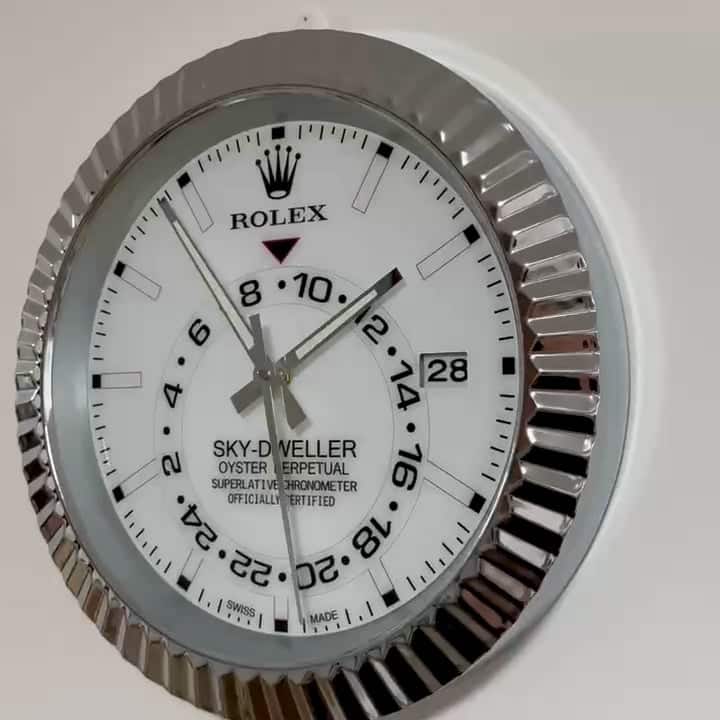 Close-up of a Rolex Sky-Dweller wall clock with a white face, silver fluted bezel, black hour markers, and date display showing the 28th.