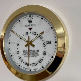 Close-up of a Rolex Sky-Dweller Oyster Perpetual wall clock with a white dial, gold hour, minute, and second hands, a gold bezel, and a date display showing the 28th, mounted on a plain light-colored wall.