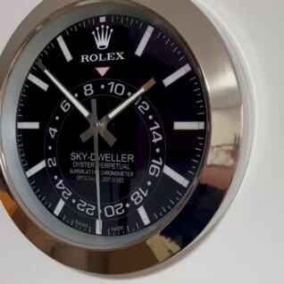 Close-up of a Rolex Sky-Dweller wall clock with a black dial, white hour markers, and a silver frame showing the time as 11:55:30.