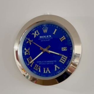 Round Rolex wall clock with a blue face, silver Roman numerals, and silver hands showing the time as 8:17:39, with a date window displaying 29 and a silver metal frame mounted on a white wall.