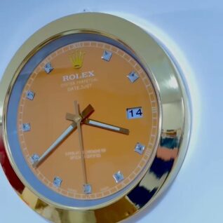 Close-up of a gold-framed Rolex Oyster Perpetual Datejust wall clock with an orange face, diamond-shaped hour markers, and showing the date as the 14th.