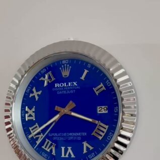 Close-up of a blue Rolex Oyster Perpetual Datejust watch face with silver Roman numerals, a date display showing 29, and a silver fluted bezel, mounted on a light grey wall.