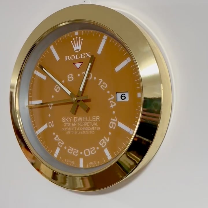 Close-up of a gold-framed Rolex Sky-Dweller wall clock with an orange dial showing the time 12:52:44 and displaying the date as the 6th.
