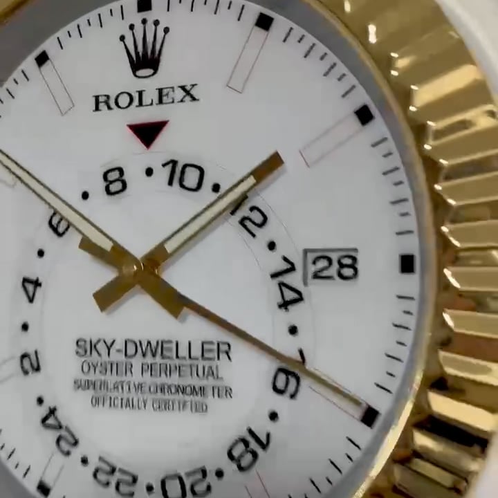 Close-up of a white Rolex Sky-Dweller watch dial showing gold hour, minute, and second hands, a date window at 3 o'clock displaying 28, and a 24-hour display with black numerals and markers, encased in a gold fluted bezel.