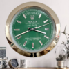 Close-up of a Rolex wall clock with a green face, silver hour markers, and a gold rim, showing the day as terça-feira and the date as 26, mounted on a white wall above a wooden surface with small decorative items including a small clock, a mug, and a glass jar with plants.