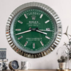 Wall clock designed to resemble a Rolex Oyster Perpetual Day-Date watch with a green face, silver hour markers, day display at the top showing terça-feira, date display showing 26, and silver edged frame, mounted on a white wall above a wooden surface with a small round clock, a decorative mug, and a glass-covered plant arrangement.