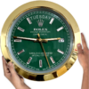 Hands holding a round Rolex wall clock with a green face, gold frame, and diamond-shaped hour markers, showing Dinsdag and the date 27 at 3 o'clock position.