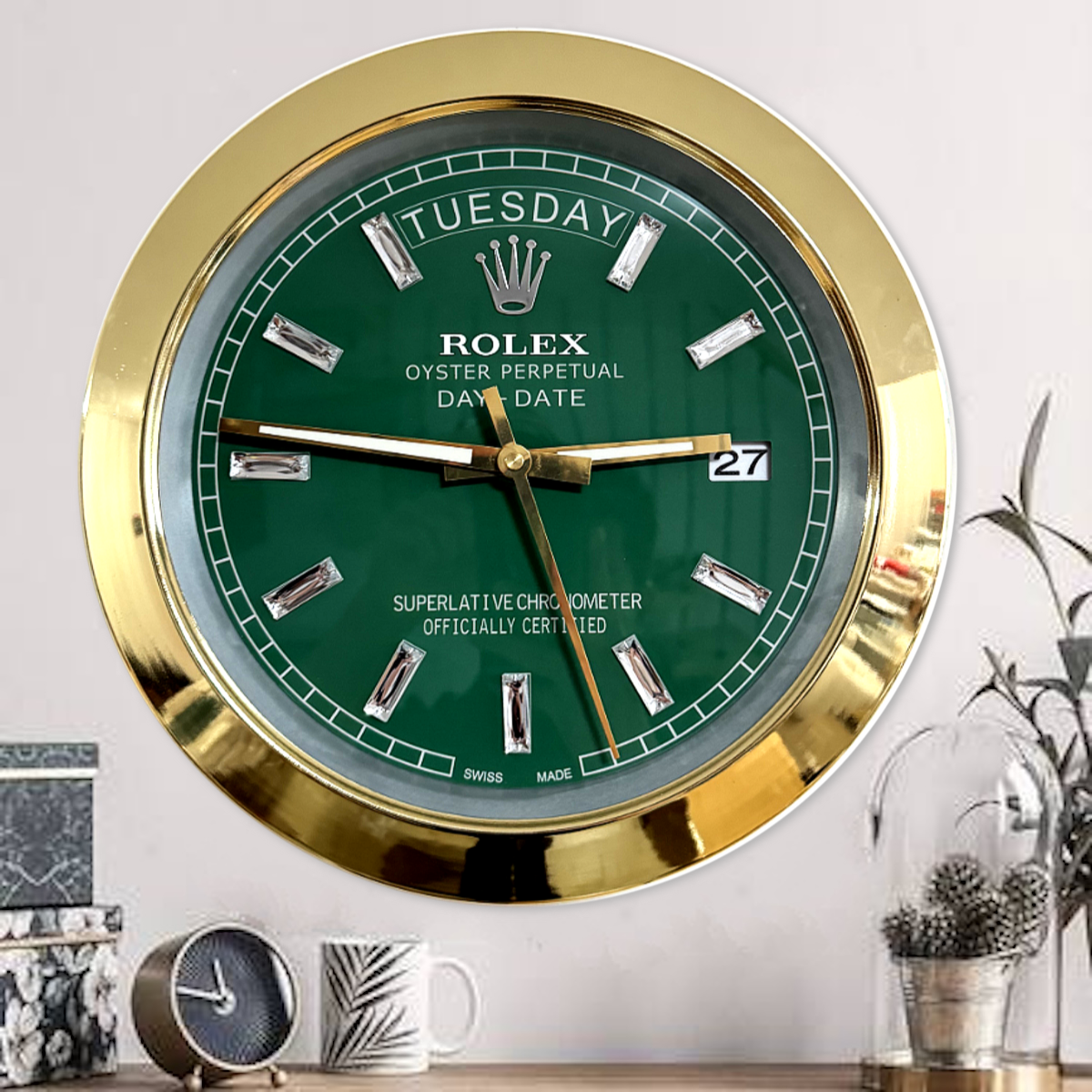 Wall-mounted Rolex clock with a green face, gold frame, and diamond-shaped hour markers showing time as 3:46:30 and displaying the day as Dinsdag and the date as 27, above a wooden surface with a small round clock, a leaf-patterned mug, floral boxes, and a plant in a glass dome on the right