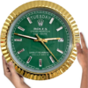 Hands holding a large Rolex clock with a gold fluted bezel, green dial displaying the day 'Dinsdag' at 12 o'clock and date '27' at 3 o'clock, with gold hour, minute, and second hands and rectangular hour markers.
