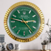 Wall clock styled like a Rolex watch face with a green dial, gold fluted bezel, and diamond hour markers, showing Dinsdag the 27th, mounted on a white wall above a wooden surface with a small round clock, a mug, and a glass cloche with dried flowers.