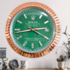 Wall clock styled like a Rolex Oyster Perpetual Day-Date watch with a green face, rose gold bezel, and diamond-shaped hour markers showing Tuesday the 26th, placed above a wooden surface with a small clock, patterned mug, and decorative plants.