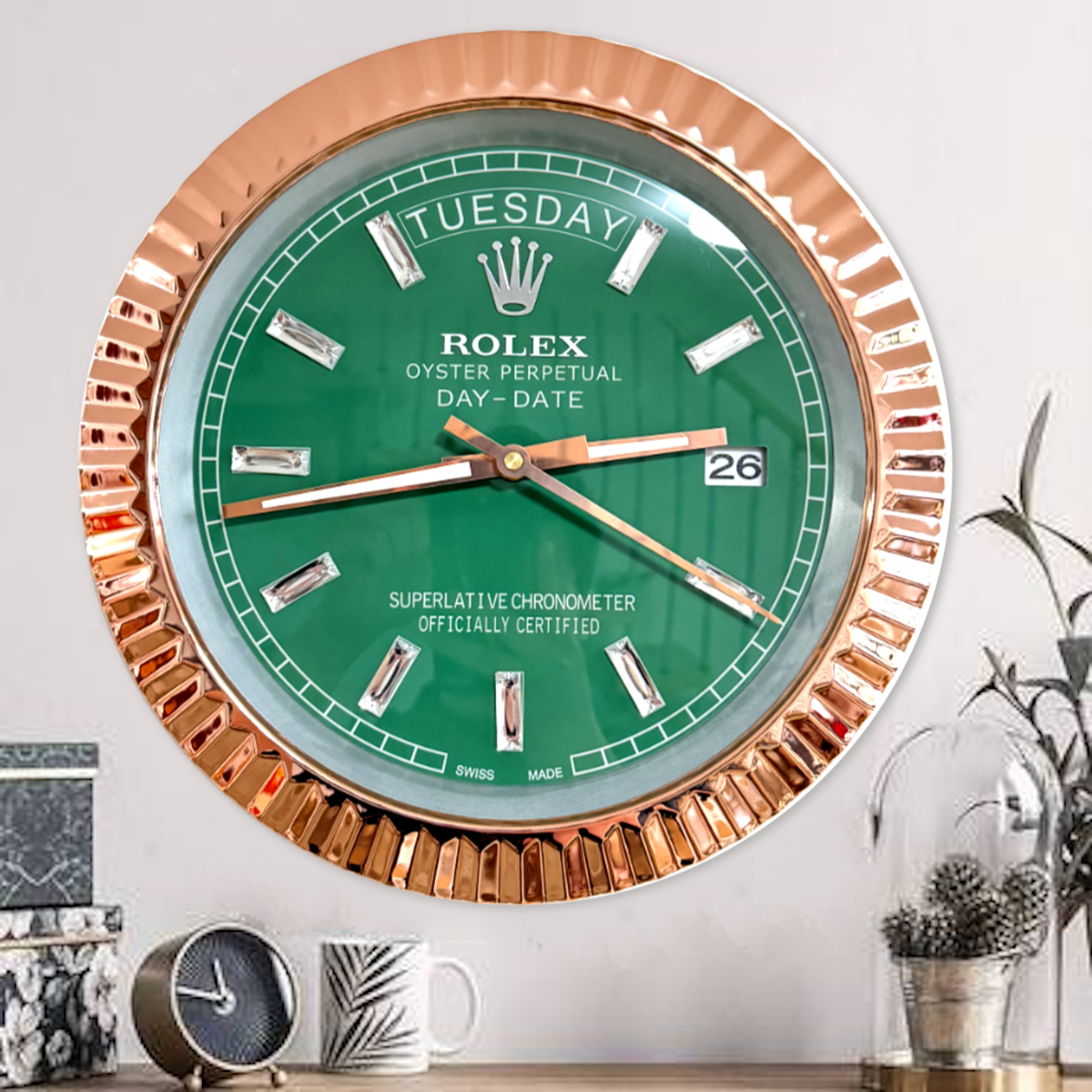 Wall clock styled like a Rolex Oyster Perpetual Day-Date watch with a green face, rose gold bezel, and diamond-shaped hour markers showing Tuesday the 26th, placed above a wooden surface with a small clock, patterned mug, and decorative plants.