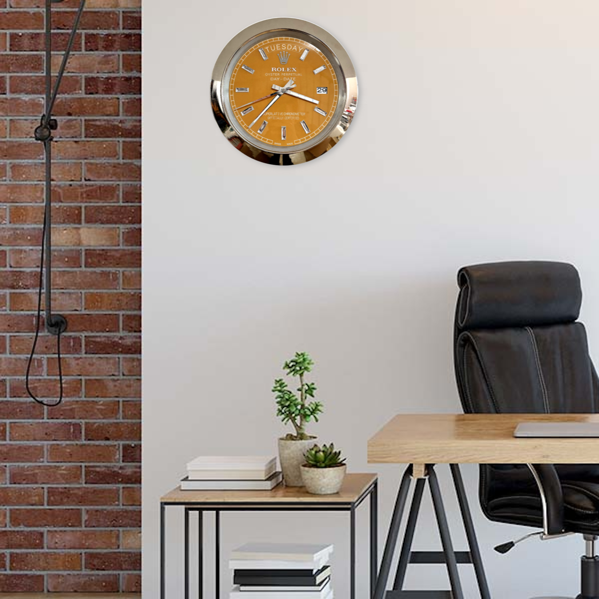 Modern office interior with a wooden desk, black leather office chair, small side table with potted plants and books, exposed brick wall on the left, and a wall clock with an orange Rolex Day-Date dial mounted on a plain light-colored wall.