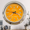 Large orange Rolex Oyster Perpetual Day-Date wall clock with silver accents, showing tirsdag and the 29th, mounted on a light wall above a table with a black clock, a white mug with leaf design, a black box with floral print, and a glass-covered plant pot containing cacti.