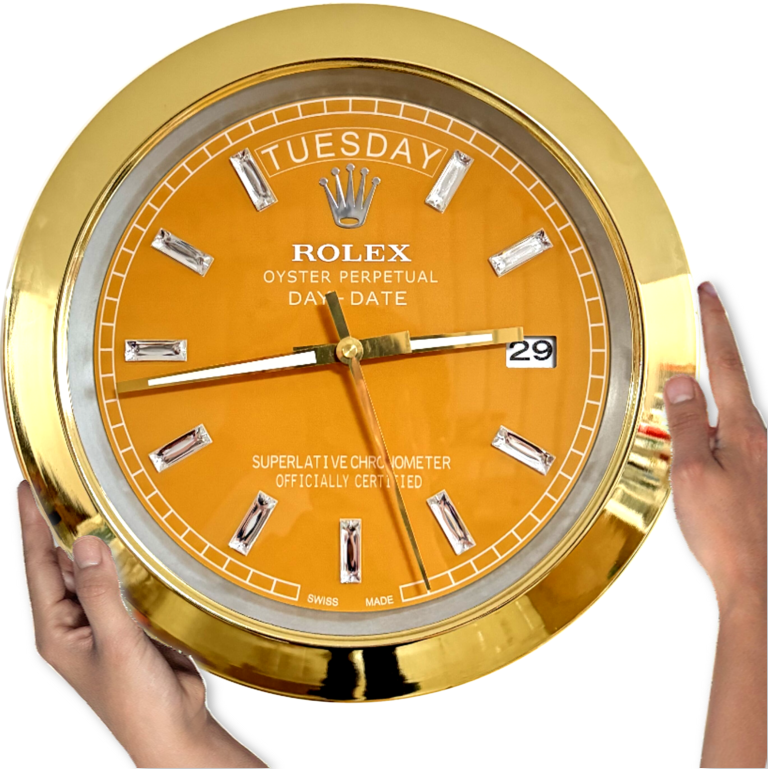 Close-up of a gold-framed Rolex wall clock with an orange face displaying the day 'tirsdag' and date '29', held by two hands on either side.
