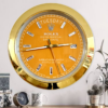 Gold-framed orange Rolex wall clock showing the day as tirsdag and the date as 29, mounted on a light grey wall above a wooden surface with a small clock, a white mug with a palm leaf design, and decorative plants in glass domes.