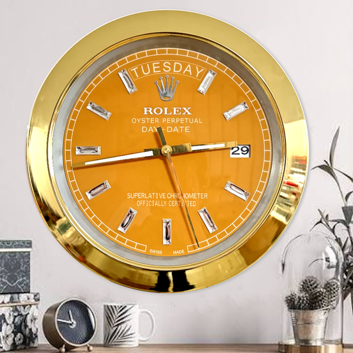 Gold-framed orange Rolex wall clock showing the day as tirsdag and the date as 29, mounted on a light grey wall above a wooden surface with a small clock, a white mug with a palm leaf design, and decorative plants in glass domes.