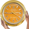 Close-up of a large Rolex Oyster Perpetual Day-Date wall clock with a gold frame and orange face, held by two hands, showing the day as Tuesday, the date as 29, and the time as approximately 3:44.
