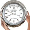 A close-up of a large, round Rolex Oyster Perpetual Day-Date wall clock with a white face, silver fluted bezel, silver hour markers, and hands, showing Salı and the date 17, held by two hands against a white background.