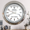 Silver-framed Rolex Oyster Perpetual Day-Date wall clock with white face, crystal hour markers, showing Salı the 17th, placed above a wooden surface with a small round clock, patterned mug, and decorative plants under a glass dome.