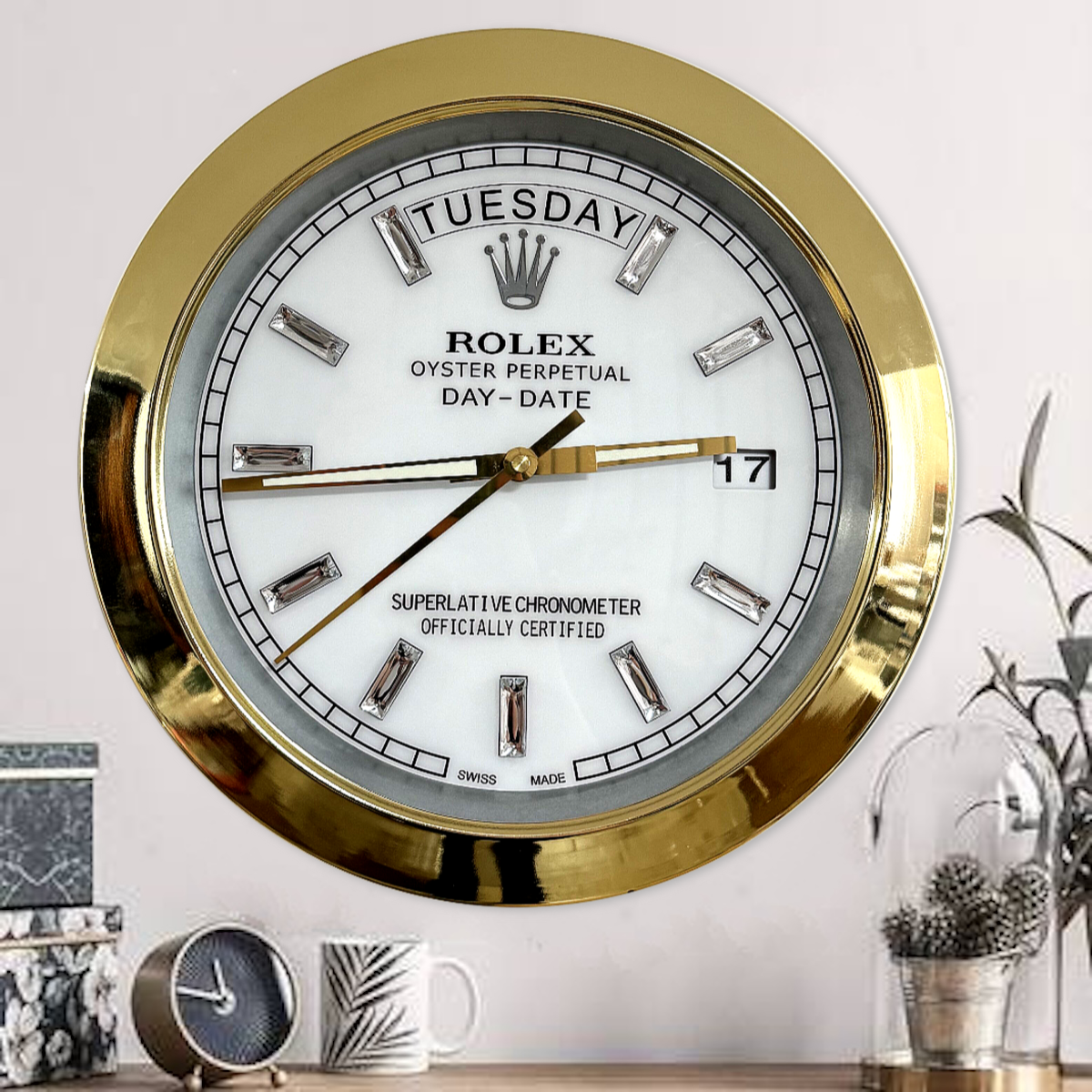 Close-up of a gold-framed Rolex Oyster Perpetual wall clock displaying the day as Dinsdag and the date as 17, with a white face and crystal hour markers, placed above a wooden surface with a small round clock, a leaf-patterned mug, and decorations including a glass dome with plants in the background.
