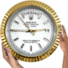 Hands holding a round Rolex wall clock with a gold fluted bezel, white dial, silver hour markers, displaying Salı and the date 17, with gold hour and minute hands and a gold second hand.