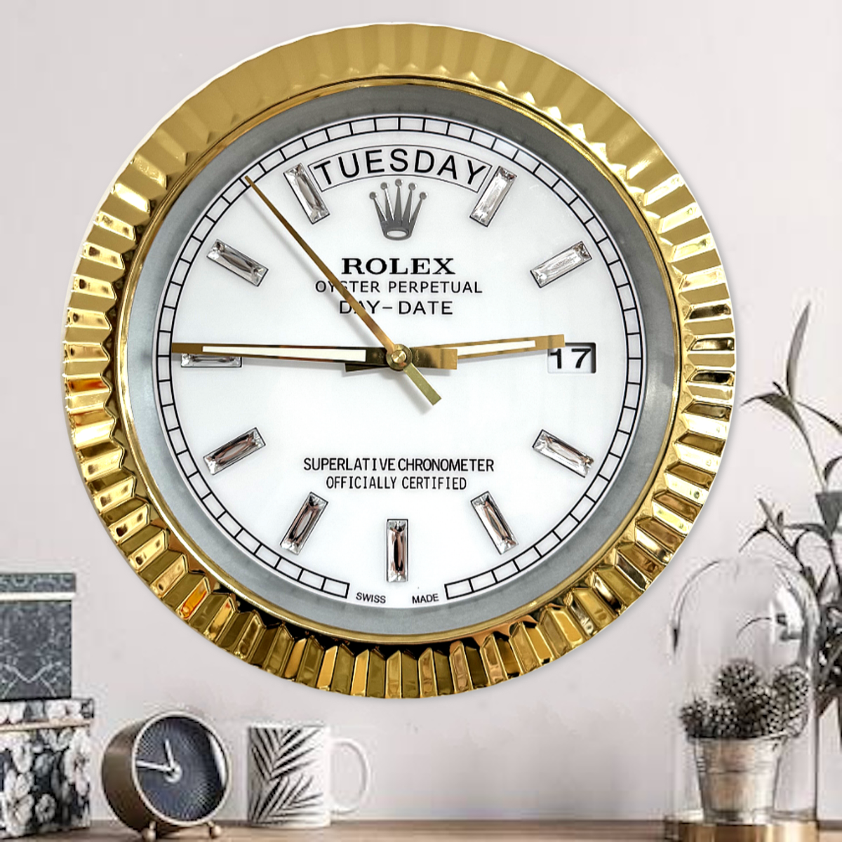 Wall clock designed to resemble a Rolex Oyster Perpetual Day-Date watch with a gold frame, white face, and diamond-shaped hour markers, showing Salı the 17th, mounted on a light-coloured wall above a wooden surface with a small clock, mug, and decorative plant nearby.