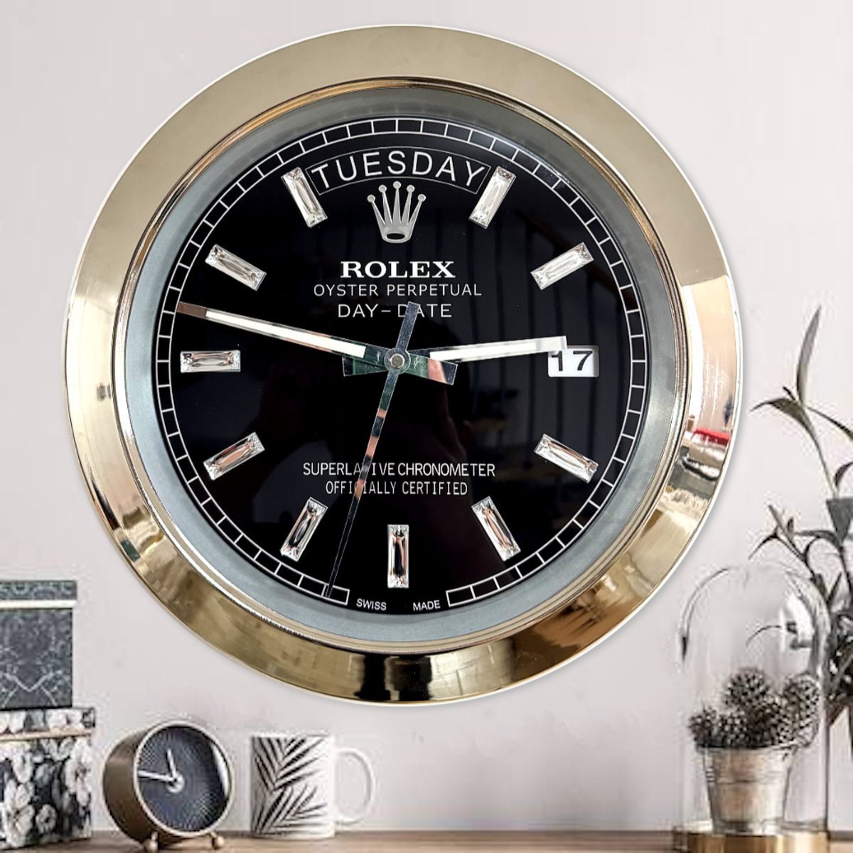 Close-up of a wall clock styled like a Rolex Oyster Perpetual Day-Date watch with a gold frame, black face, diamond-shaped hour markers, and showing terça-feira the 17th, placed above a wooden surface with a small round black clock, a mug with a leaf design, and a glass dome covering a plant with pinecones inside.