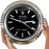 Large round black Rolex wall clock with silver fluted bezel displaying Dinsdag and date 17, held by two hands on either side