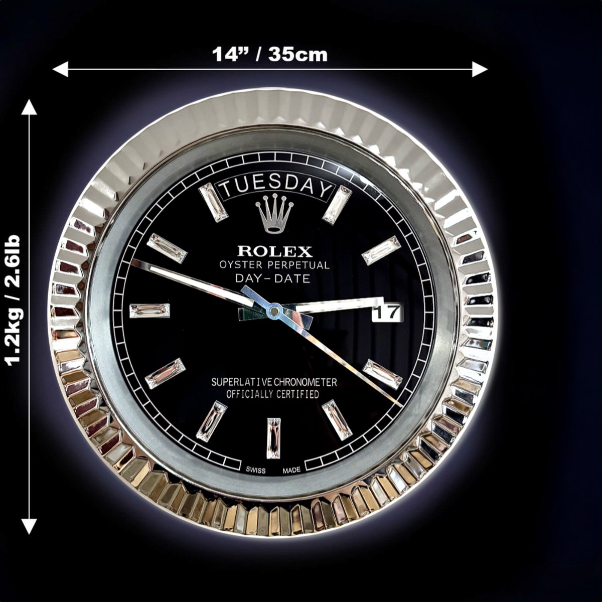 Close-up of a round Rolex Oyster Perpetual Day-Date wall clock with a black face, silver hour markers, and silver bezel, showing the day as Dinsdag and the date as 17, with dimensions 14 inches (35 cm) diameter, weighing 1.2 kg (2.6 lb) indicated by white measurement arrows on a dark background.