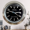 Wall clock designed to resemble a Rolex Oyster Perpetual Day-Date watch face with black dial, silver hour markers, and a fluted silver bezel, showing Dinsdag and the date as 17, hanging on a white wall above a wooden surface with a small round clock, a white mug with leaf design, and a glass container with decorative pine cones and a plant.