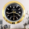 Wall clock with a gold frame and black face designed to resemble a Rolex Oyster Perpetual Day-Date watch, showing the day as Tuesday and the date as 17, mounted on a light wall above a wooden surface with decorative items including a small clock, a mug, and plants.