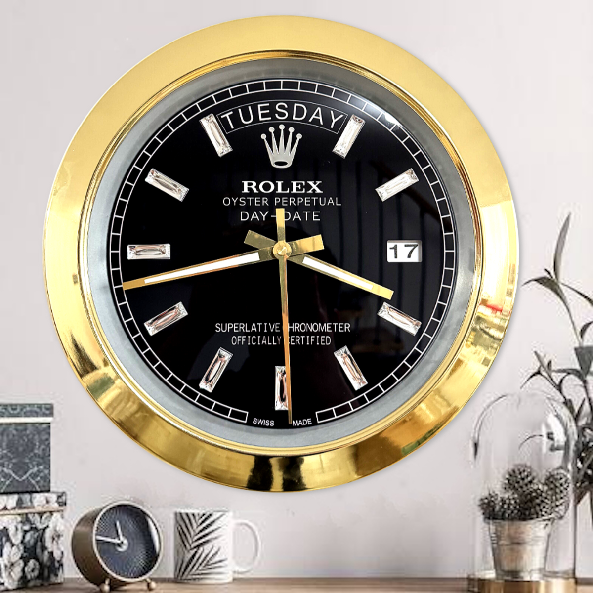 Wall clock with a gold frame and black face designed to resemble a Rolex Oyster Perpetual Day-Date watch, showing the day as Tuesday and the date as 17, mounted on a light wall above a wooden surface with decorative items including a small clock, a mug, and plants.