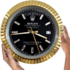 Close-up of a black and gold Rolex Oyster Perpetual Day-Date wall clock showing the time as 9:18:29 with day displayed as Martedì and date as 17, held by two hands on either side against a white background.