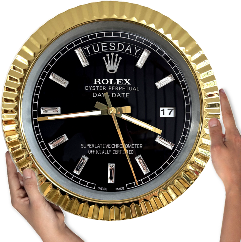 Close-up of a black and gold Rolex Oyster Perpetual Day-Date wall clock showing the time as 9:18:29 with day displayed as Martedì and date as 17, held by two hands on either side against a white background.