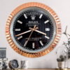 Wall clock designed like a Rolex Oyster Perpetual Day-Date watch with a black face, gold fluted bezel, diamond hour markers, and day-date display showing terça-feira and the 17th, mounted on a beige wall above a wooden surface with decorative items.