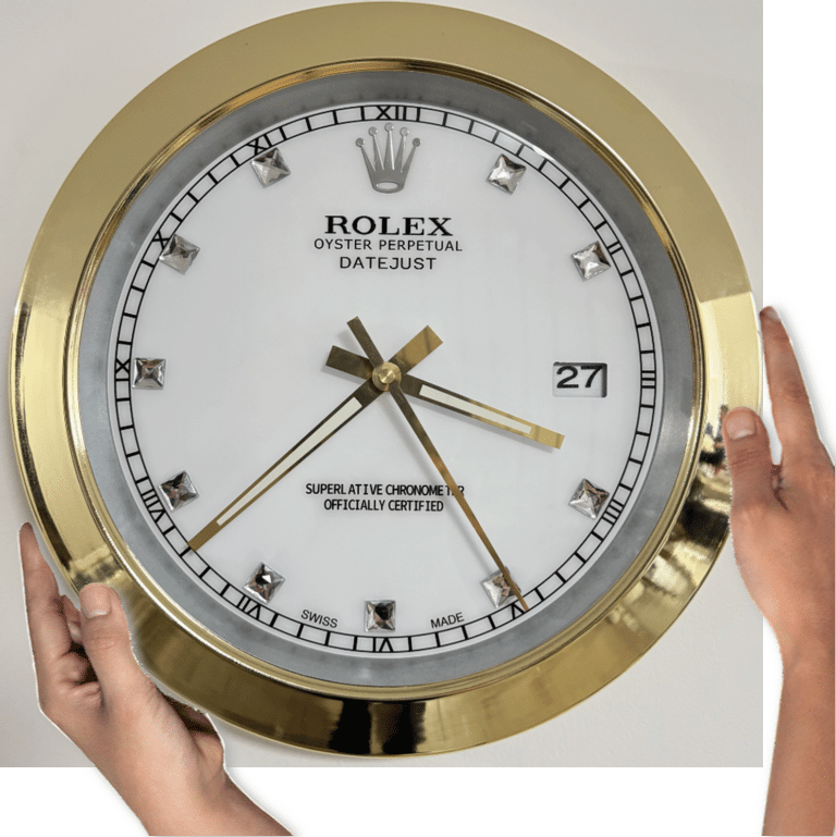 Close-up of hands holding a gold-rimmed Rolex wall clock with a white face showing the time as 3:23, featuring Roman numerals and diamond-like hour markers, with the date displayed as 27.