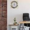 Office corner with a black leather chair, wooden desk, small plant on a wooden side table, stack of books, and a wall clock designed to resemble a Rolex watch hanging on a white wall next to a red brick wall.
