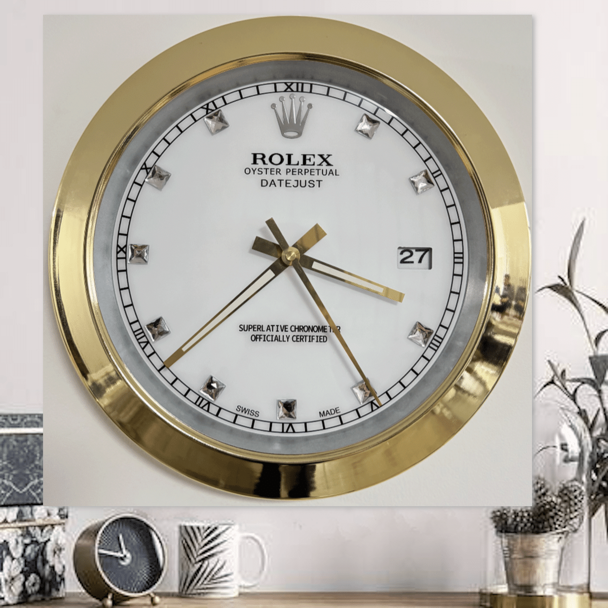 Gold-rimmed Rolex wall clock with diamond hour markers, white face, date window showing 27, and three gold hands indicating time around 3:37:25, mounted on a white wall above a desk with a small clock, mug, and plants.